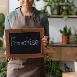 Woman holding a chalkboard with "Franchise" in a plant-filled setting.