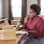 Home office workspace with woman working on laptop and packages on table.